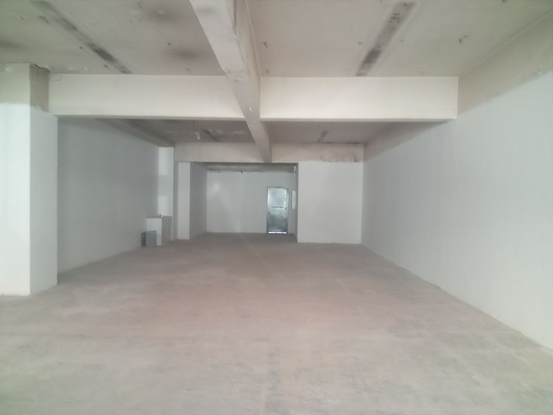 An open-plan retail space in shell condition, featuring exposed concrete flooring, structural beams, and a rear access door.