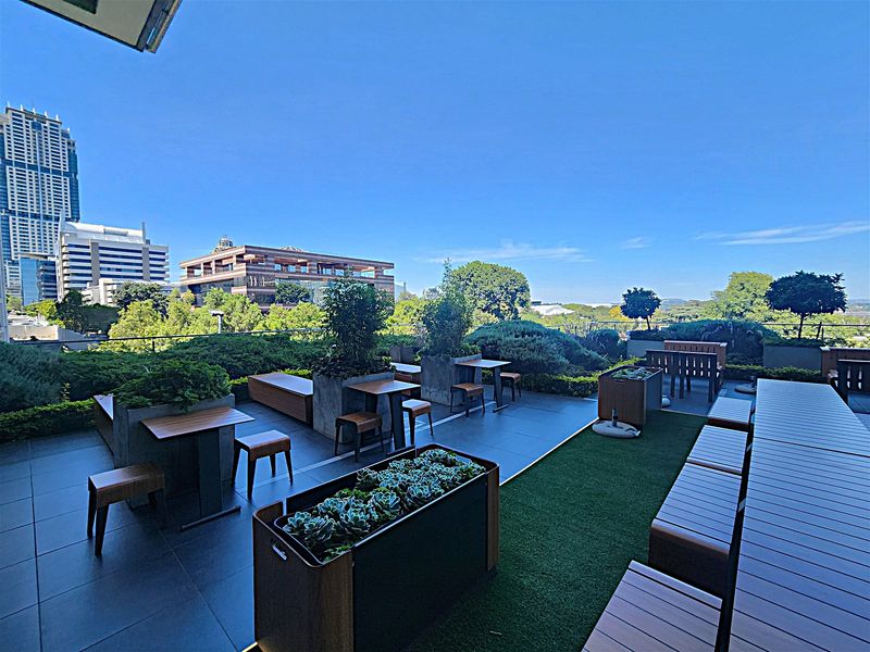 A spacious, well-maintained outdoor terrace featuring tiled flooring, artificial turf sections, built-in planters, and a glass balustrade with expansive city views.