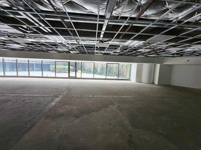 An expansive, unfinished office space featuring an exposed ceiling grid, concrete flooring, and large floor-to-ceiling windows overlooking an outdoor area.