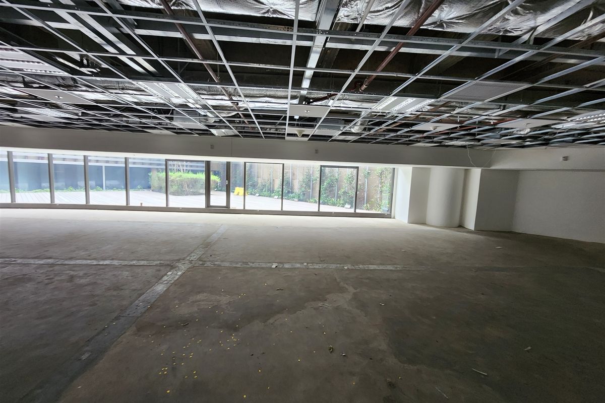 An expansive, unfinished office space featuring an exposed ceiling grid, concrete flooring, and large floor-to-ceiling windows overlooking an outdoor area.