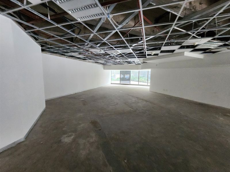 An empty, unfinished office space featuring an exposed ceiling grid, concrete flooring, and large windows providing natural light.
