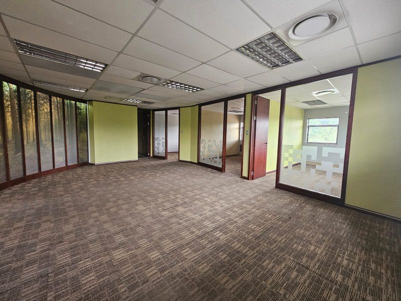 An open-plan office space featuring carpeted flooring, suspended ceilings with recessed lighting, and glass partition walls creating multiple private office areas.