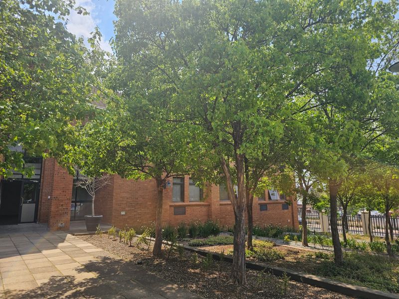 A well-maintained brick commercial building exterior featuring a paved entrance walkway, mature trees, and landscaped garden beds.