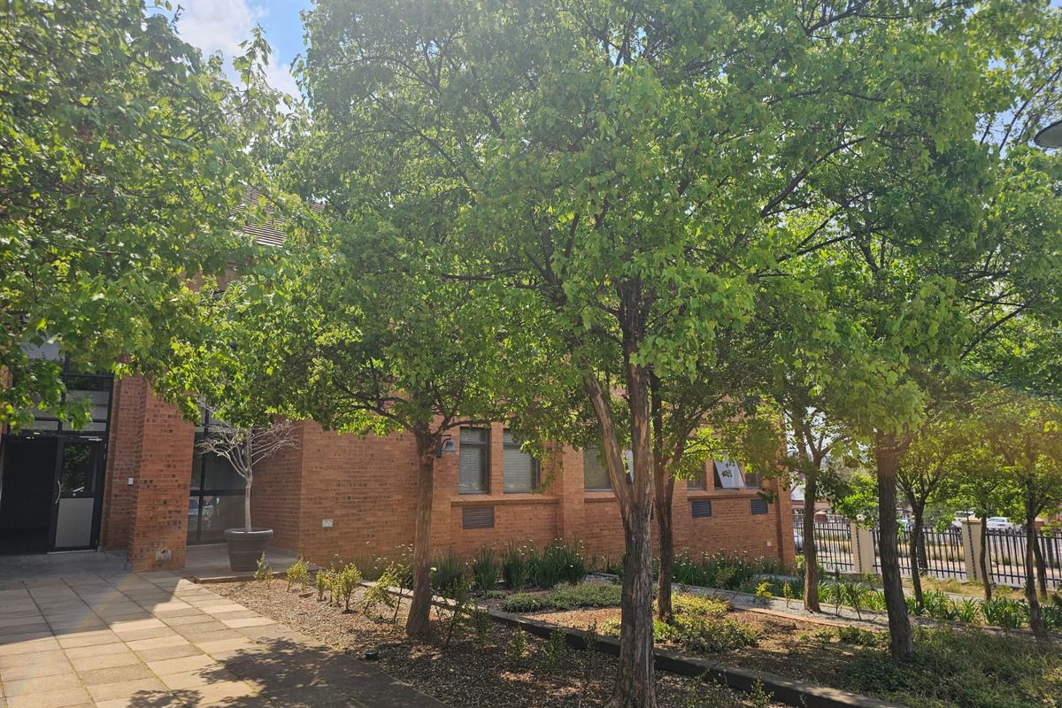 A well-maintained brick commercial building exterior featuring a paved entrance walkway, mature trees, and landscaped garden beds.