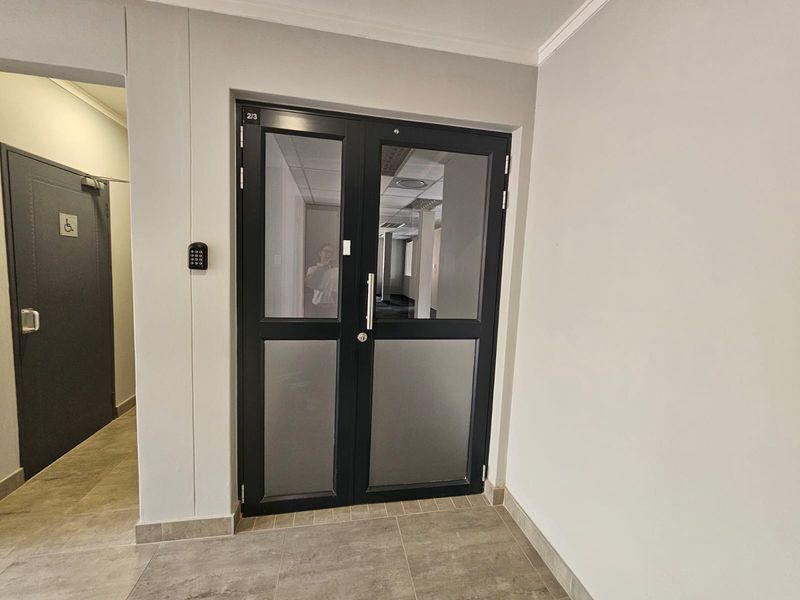A clean office entry area featuring double glass doors with a digital keypad lock and accessible signage, leading into a professional workspace.