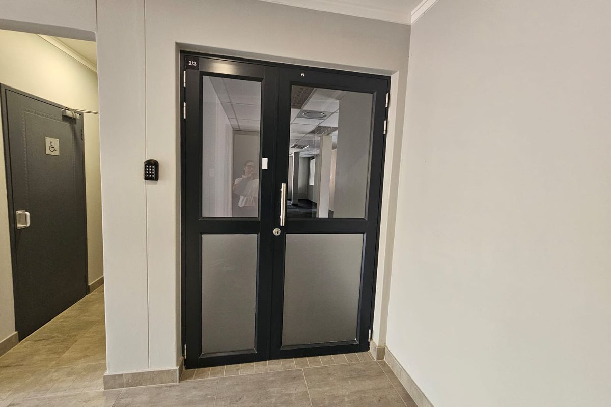 A clean office entry area featuring double glass doors with a digital keypad lock and accessible signage, leading into a professional workspace.