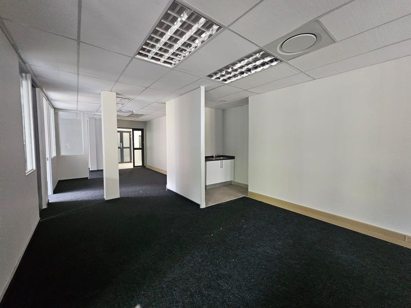 Open plan office space featuring a kitchenette, suspended ceilings with recessed lighting, and dark carpeting throughout.