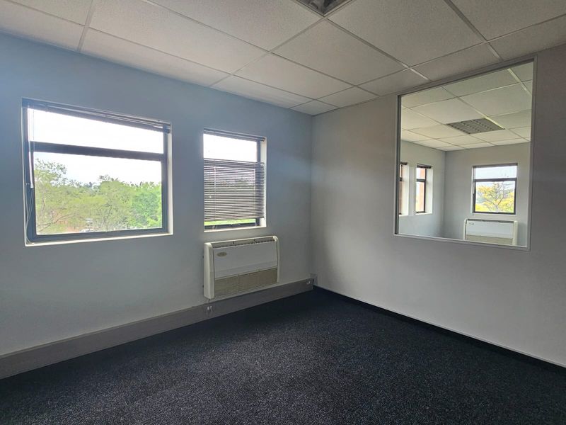 A vacant office space featuring dark carpeting, a suspended ceiling, wall-mounted air conditioning, and a large wall mirror reflecting the adjacent room.