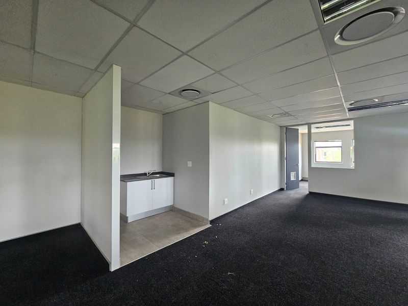 An open-plan office space featuring a small kitchenette area, suspended ceilings, recessed lighting, and dark carpeting.