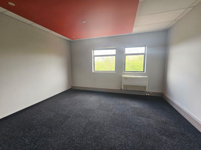 A vacant office space featuring dark carpet flooring, a suspended ceiling with a red accent panel, and a wall-mounted air conditioning unit.
