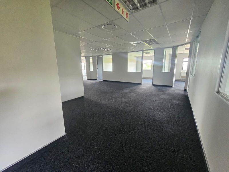 An open-plan office space featuring carpet tiles, suspended ceilings with recessed lighting, and glass partition walls creating separate work areas.