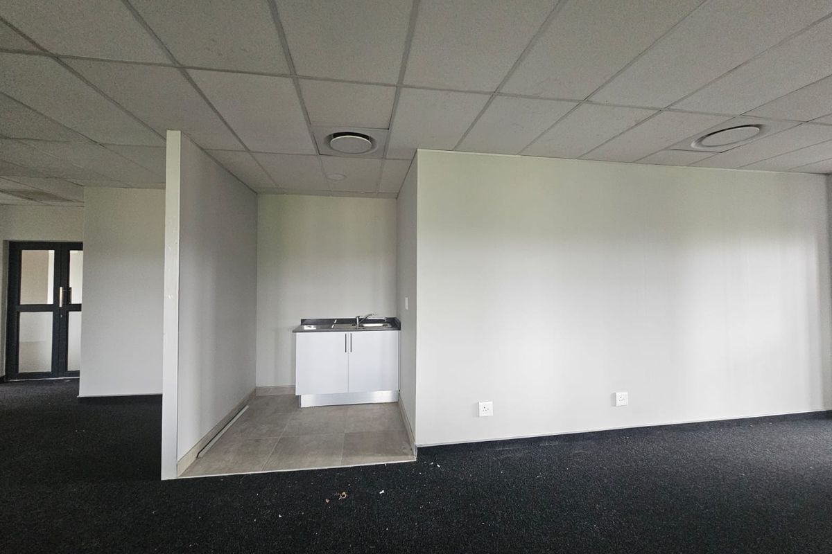 A vacant office kitchenette area featuring a small cabinet with a stainless steel sink, drop ceiling, and dark carpeting.