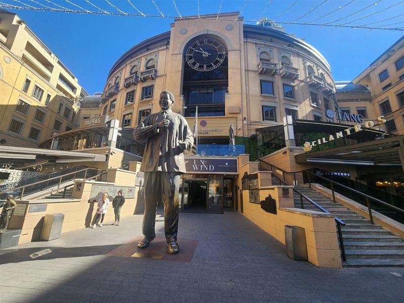 Exterior view of Nelson Mandela Square featuring a prominent statue, large clock, and surrounding commercial office and retail building architecture.