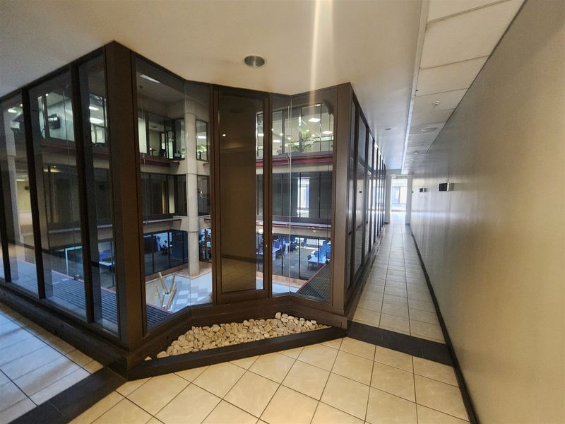 A bright office hallway featuring extensive glass partitioning overlooking an internal atrium, finished with clean tiled flooring and modern recessed lighting.