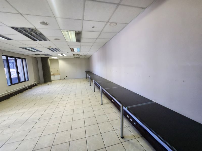 Spacious open-plan office area featuring tiled flooring, a suspended ceiling with recessed lighting, and a long built-in desk unit.