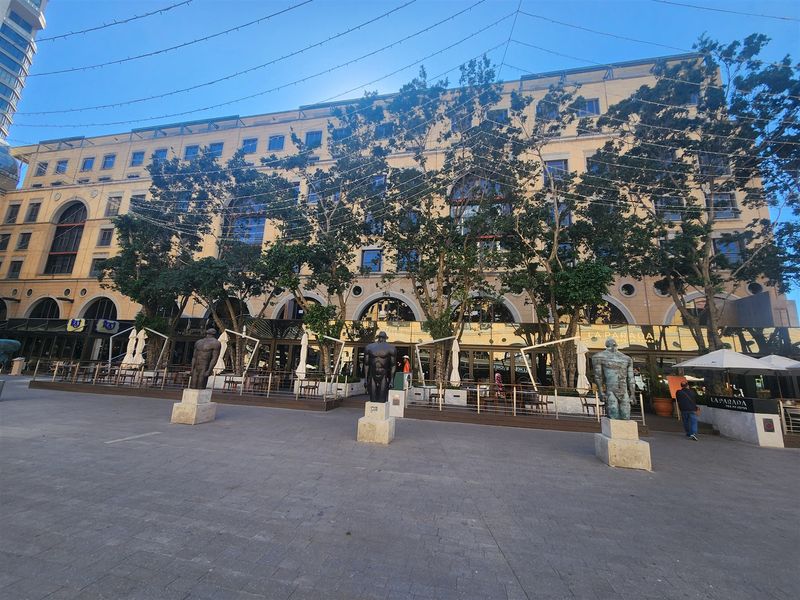 A wide-angle view of a multi-story commercial office building facade featuring a paved plaza, decorative statues, and ground-level outdoor dining areas.