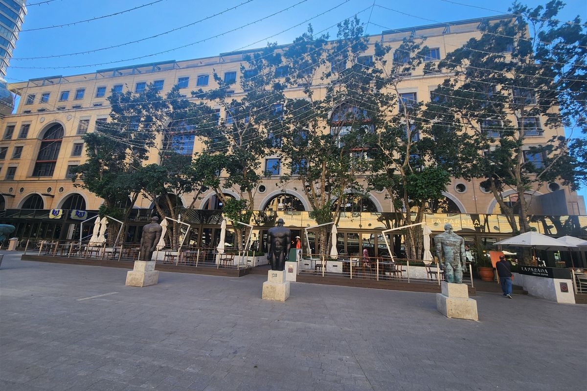 A wide-angle view of a multi-story commercial office building facade featuring a paved plaza, decorative statues, and ground-level outdoor dining areas.