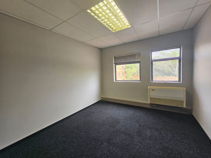 A clean, empty office space featuring carpeted flooring, a suspended ceiling with recessed lighting, and a wall-mounted air conditioning unit.