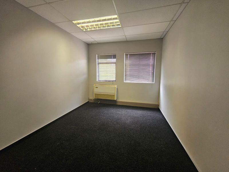 A clean, empty office space featuring carpeted flooring, a suspended ceiling with recessed lighting, and a wall-mounted air conditioning unit.