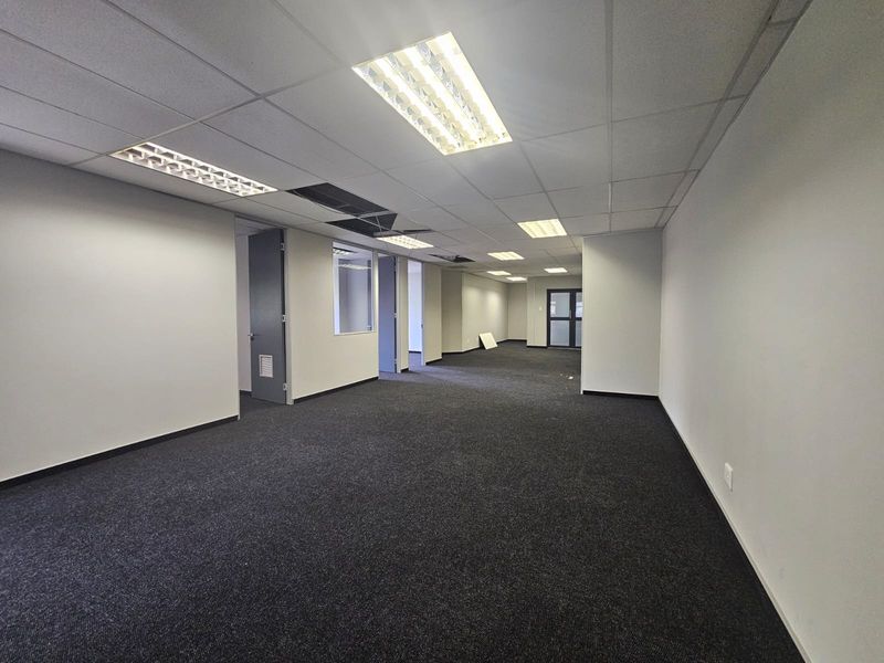 An empty, open-plan office space featuring dark carpeting, a suspended ceiling with recessed lighting, and an internal glass partition.