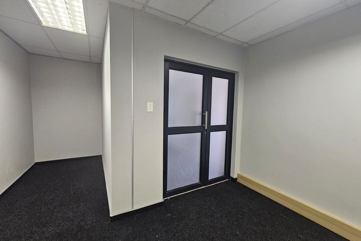 A clean, empty office space featuring dark carpeting, a suspended ceiling with recessed lighting, and a modern frosted glass double door entrance.