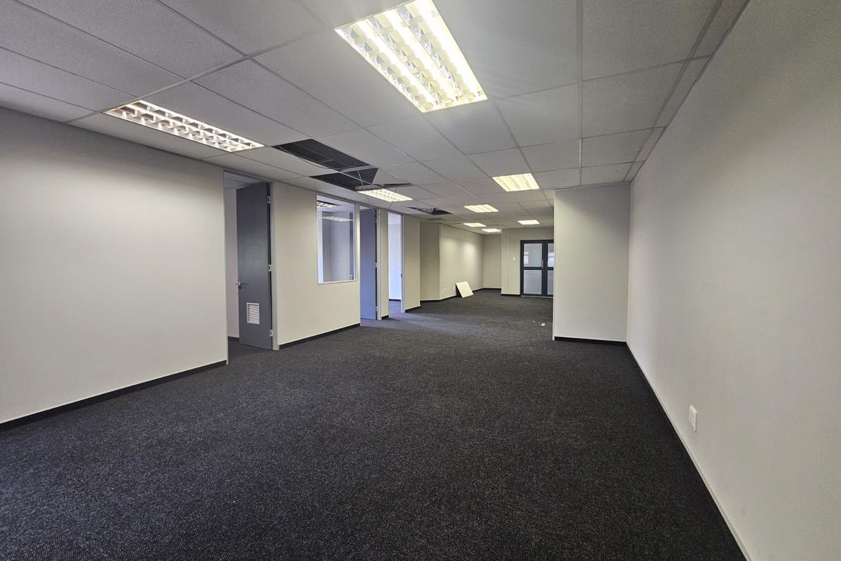 An empty, open-plan office space featuring dark carpeting, a suspended ceiling with recessed lighting, and an internal glass partition.