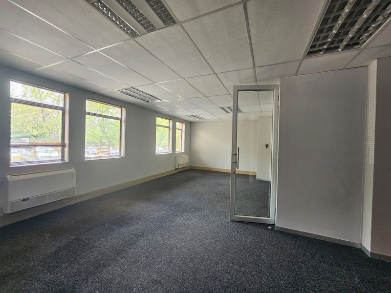 Empty office space featuring carpeted flooring, suspended ceilings with recessed lighting, wall-mounted air conditioning units, and large windows providing natural light.