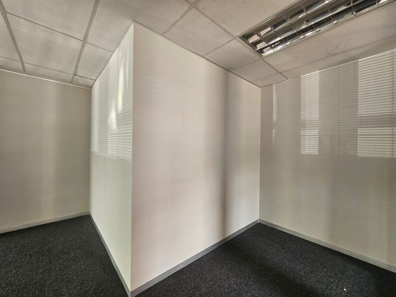 A vacant, neutral office space featuring carpet tiles, a suspended ceiling with recessed lighting, and venetian blinds on the windows.