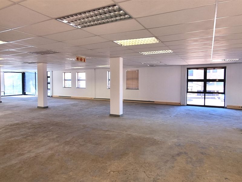 A large, open-plan office space featuring a suspended ceiling with recessed lighting, structural columns, and large windows, currently awaiting floor finishing.