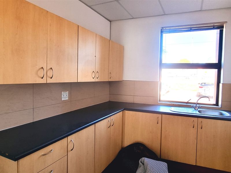 A functional office kitchenette featuring light wood-finish cabinetry, dark laminate countertops, a stainless steel sink, and a tiled backsplash.