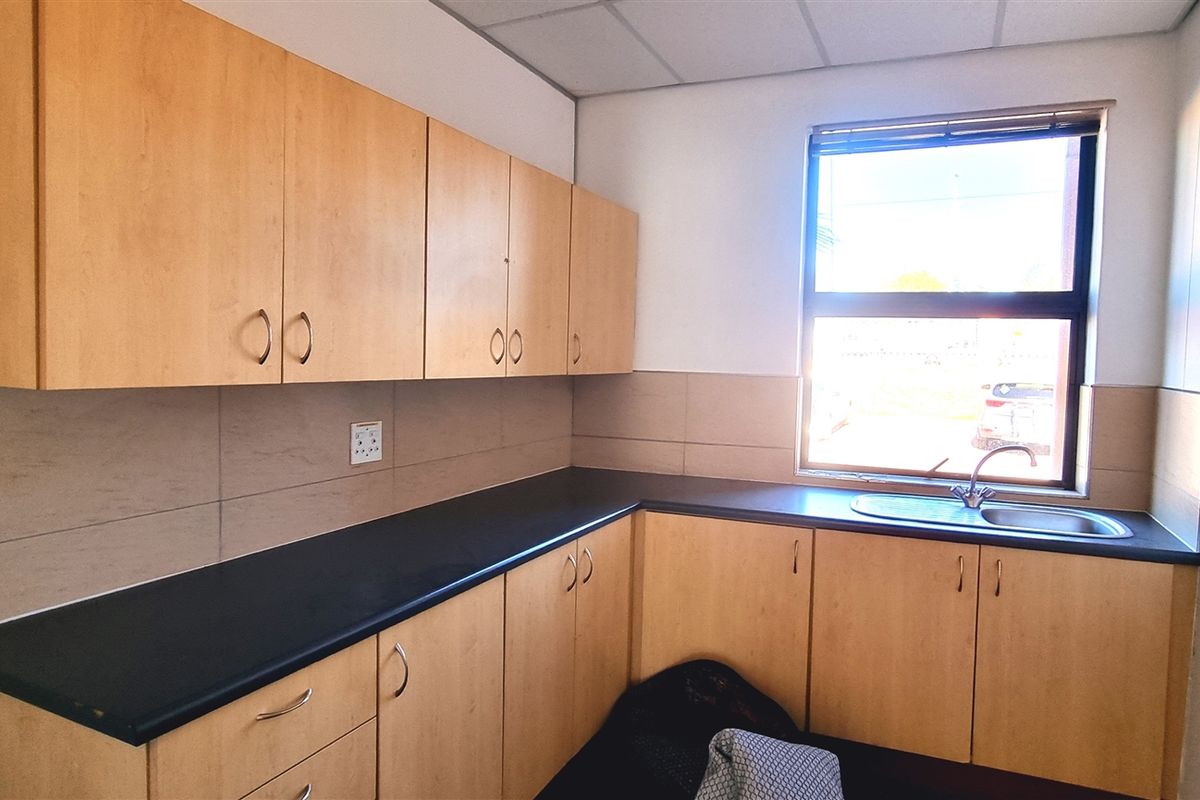 A functional office kitchenette featuring light wood-finish cabinetry, dark laminate countertops, a stainless steel sink, and a tiled backsplash.