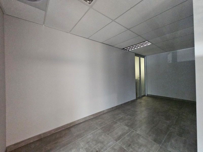 A clean, empty office space featuring modern tiled flooring, a suspended ceiling with recessed lighting, and a glass partition door.