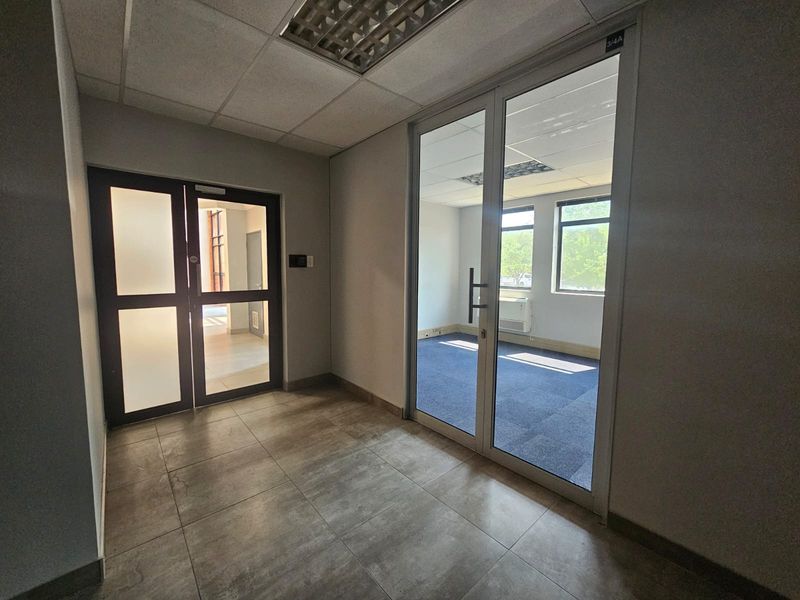 A clean, professional office hallway featuring tiled flooring, glass partition doors, and a suspended ceiling, leading into a vacant carpeted office space.