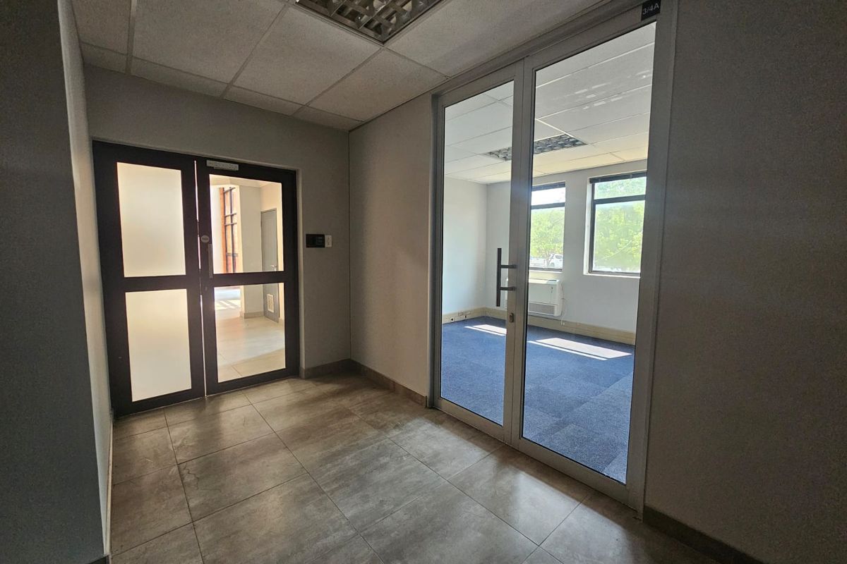 A clean, professional office hallway featuring tiled flooring, glass partition doors, and a suspended ceiling, leading into a vacant carpeted office space.