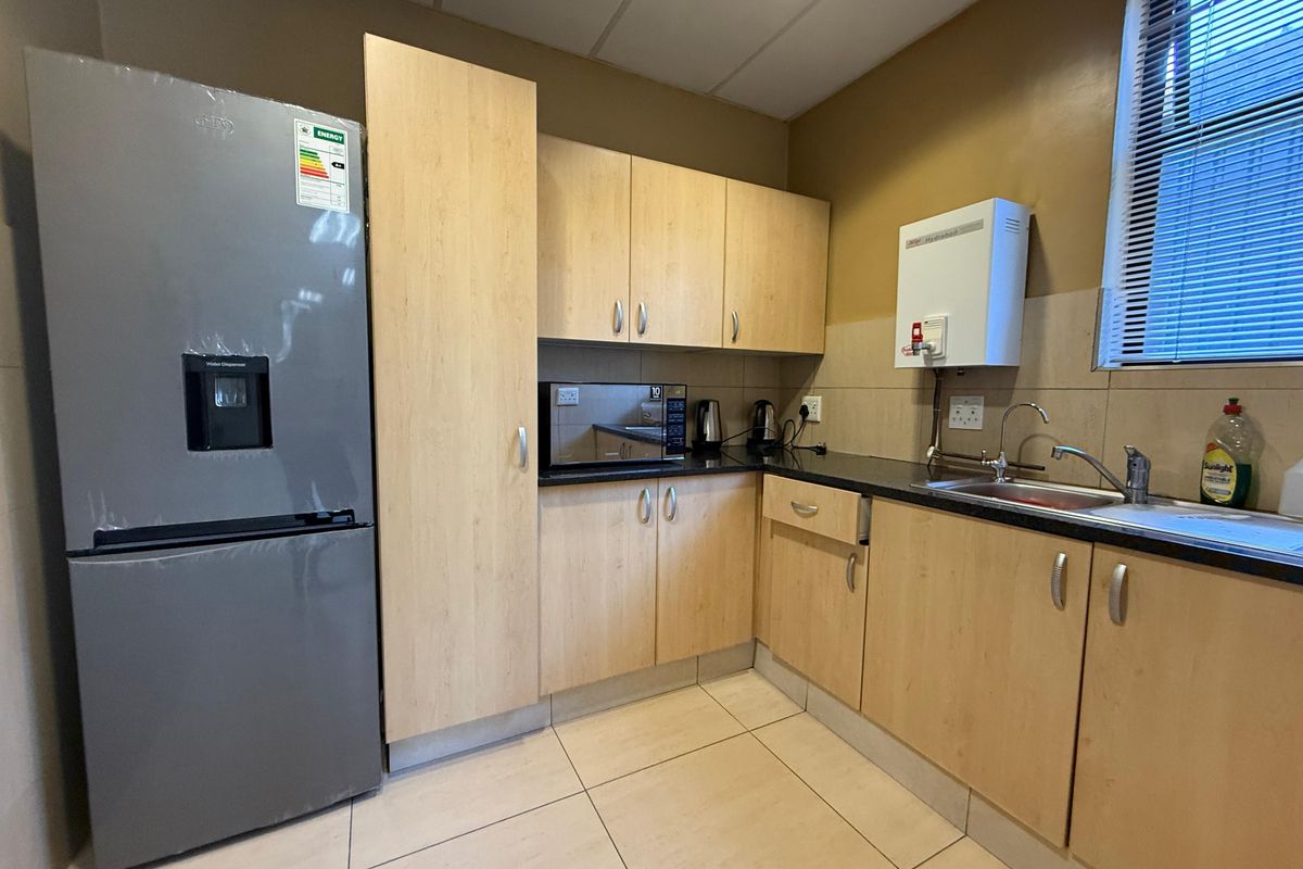 A functional office kitchenette featuring light wood cabinetry, dark countertops, a stainless steel sink, and a wall-mounted water heater.