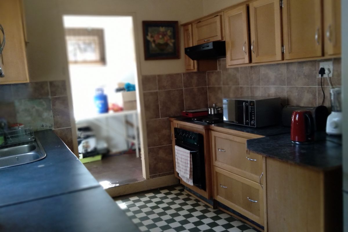 A functional kitchen featuring wooden cabinetry, laminate countertops, a built-in oven with extractor fan, and classic checkered floor tiles, leading into a secondary utility area.