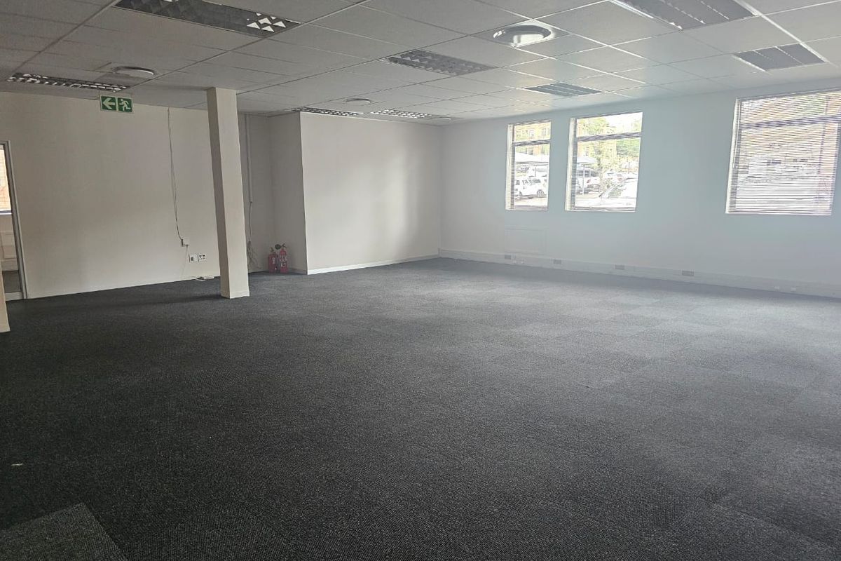 Spacious, open-plan office area featuring a suspended ceiling, recessed lighting, carpeted flooring, and large windows with venetian blinds.