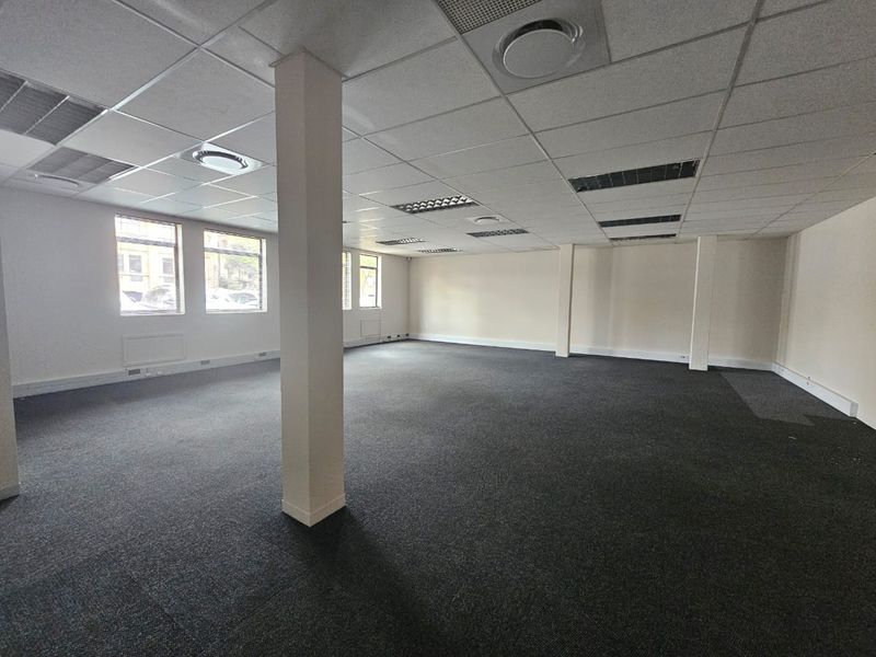 A spacious, open-plan office area featuring neutral carpeting, a suspended ceiling with recessed lighting, and large windows providing natural light.