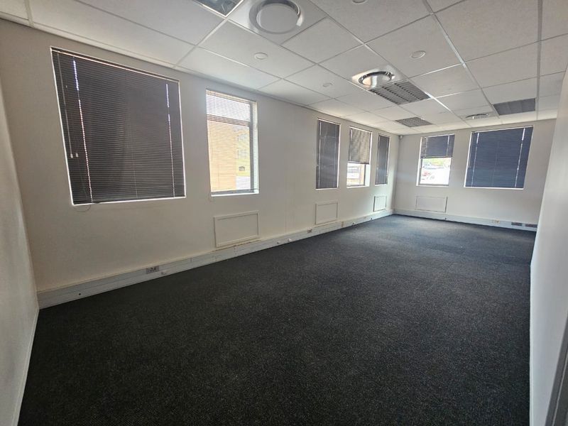 A vacant, open-plan office space featuring a suspended ceiling, recessed lighting, carpeted flooring, and multiple windows with fitted venetian blinds.