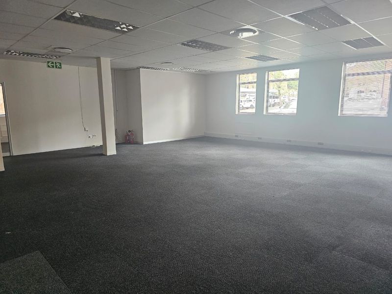 Spacious, open-plan office area featuring a suspended ceiling, recessed lighting, carpeted flooring, and large windows with venetian blinds.