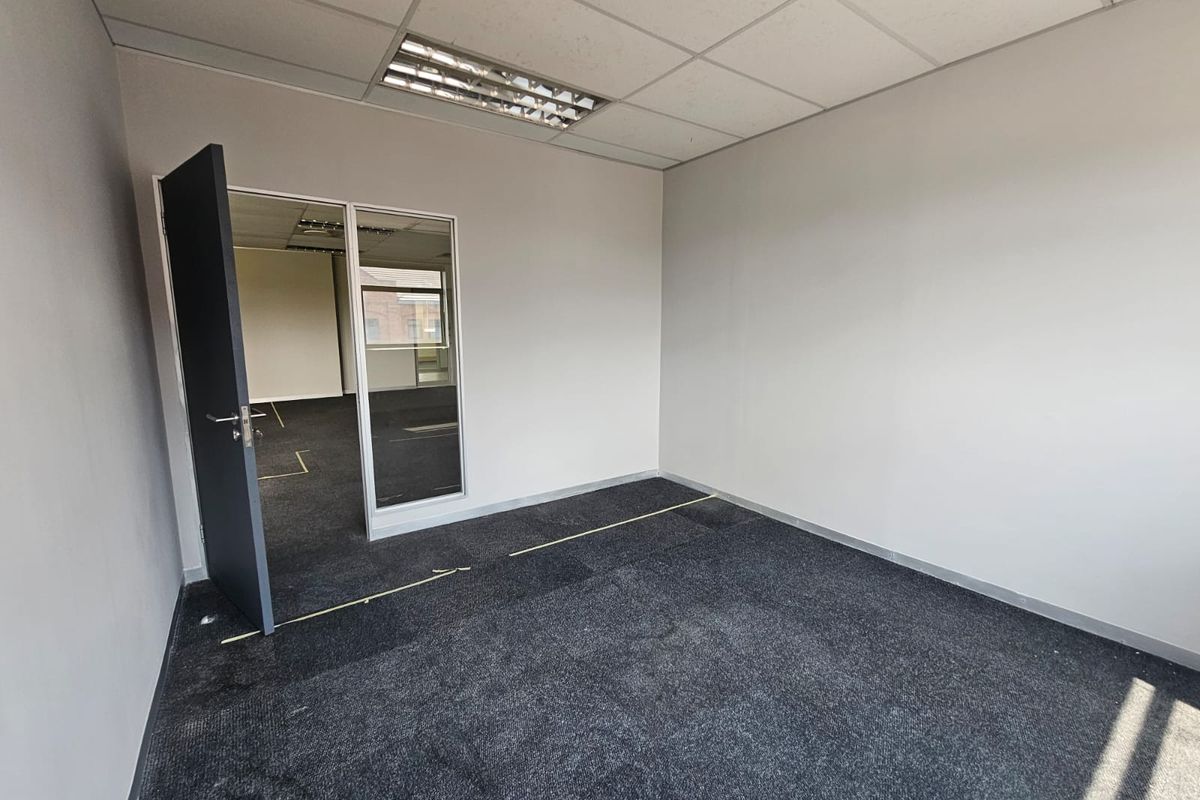 An empty office space featuring dark carpeting, a suspended ceiling with integrated lighting, and a glass partition wall.