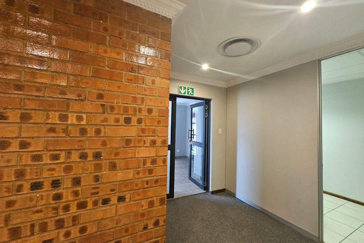 A clean office hallway featuring an exposed brick feature wall, recessed lighting, and glass partition doors leading to adjacent workspaces.