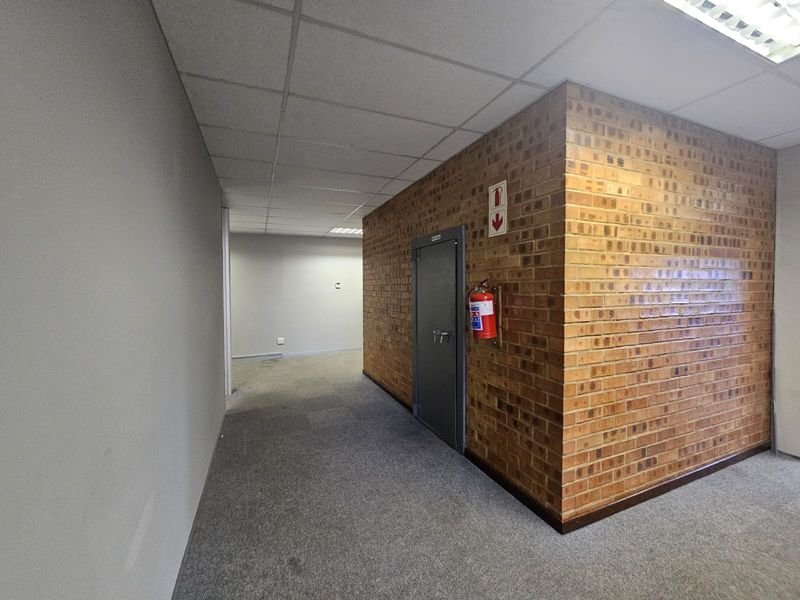 A functional office hallway featuring a secure door, exposed brick wall, suspended ceiling, and carpeted flooring.