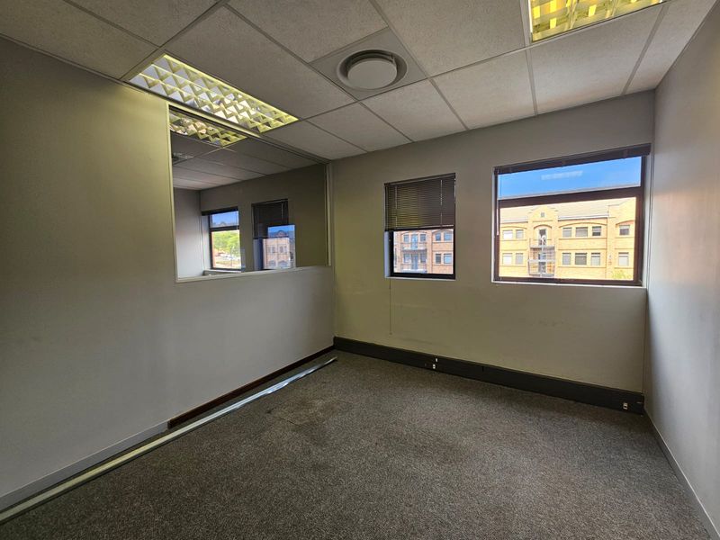 An empty office space featuring a suspended ceiling, recessed lighting, carpeted flooring, and an internal glass partition overlooking an adjacent area.