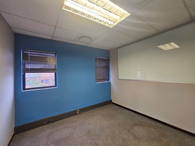 A vacant office space featuring carpeted flooring, a suspended ceiling with integrated lighting, venetian blinds, and a glass partition wall.