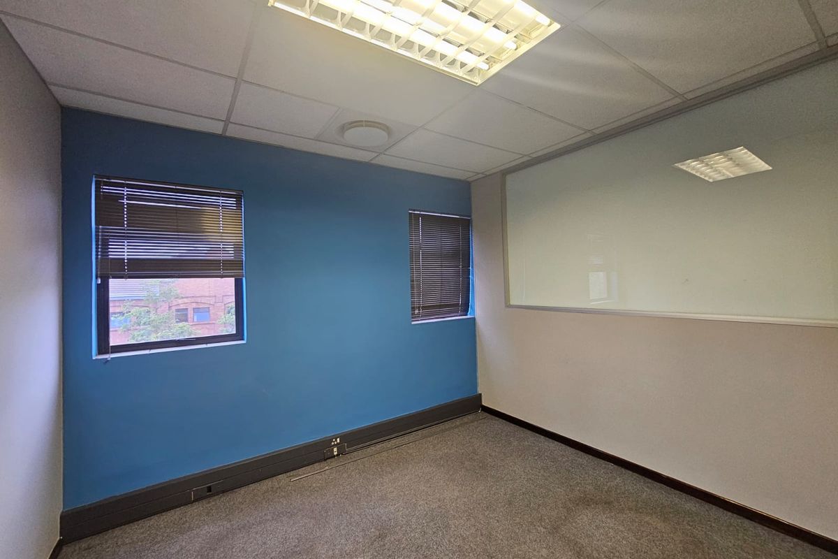 A vacant office space featuring carpeted flooring, a suspended ceiling with integrated lighting, venetian blinds, and a glass partition wall.