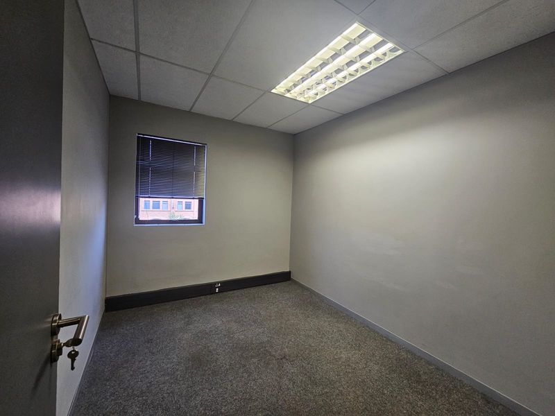 A vacant, neutral-toned office space featuring carpeted flooring, a suspended ceiling with recessed lighting, and a window with fitted venetian blinds.