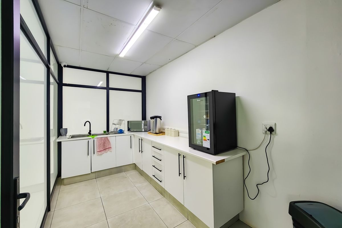 A functional office kitchenette featuring white cabinetry, a stainless steel sink, tiled flooring, and a glass partition wall.