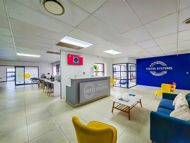 Modern office reception area featuring a branded desk, tiled flooring, suspended ceiling with recessed lighting, and glass-partitioned workspace areas.