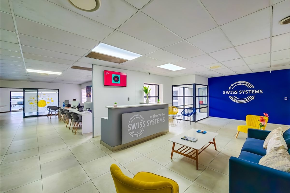 Modern office reception area featuring a branded desk, tiled flooring, suspended ceiling with recessed lighting, and glass-partitioned workspace areas.